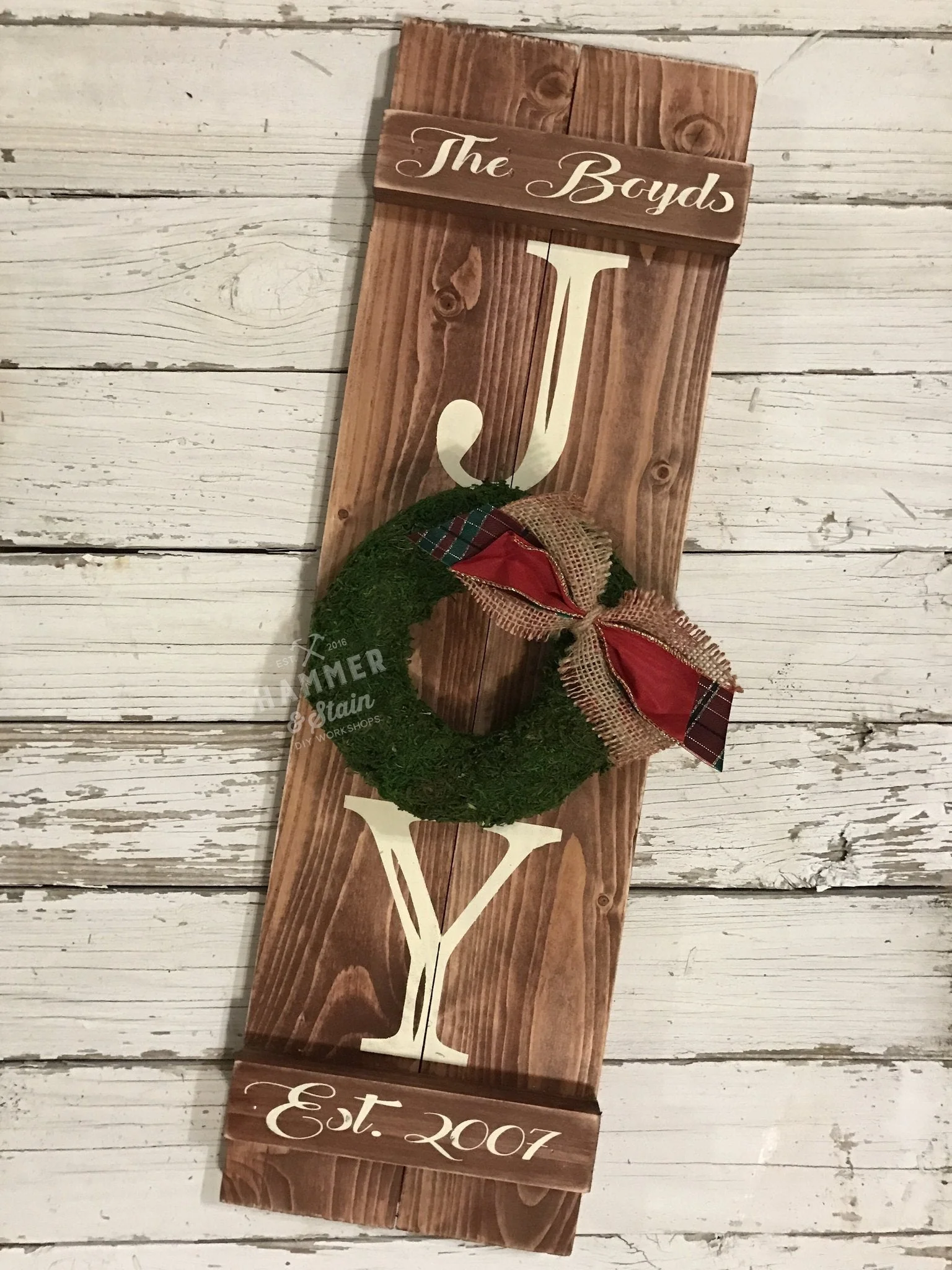Picture of a two board shutter with a family name across the top, the year at the bottom and Joy down the front with a wreath for the "o" with a rustic red, plaid and burlap bow.